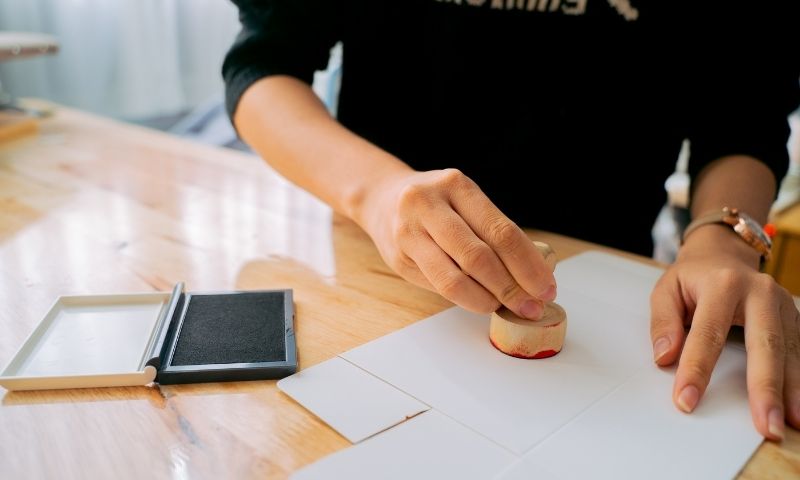 Person mit schwarzem Pulli drückt Stempel auf weißes Papier, das auf einem Holztisch liegt, links ist ein Stempelkissen
