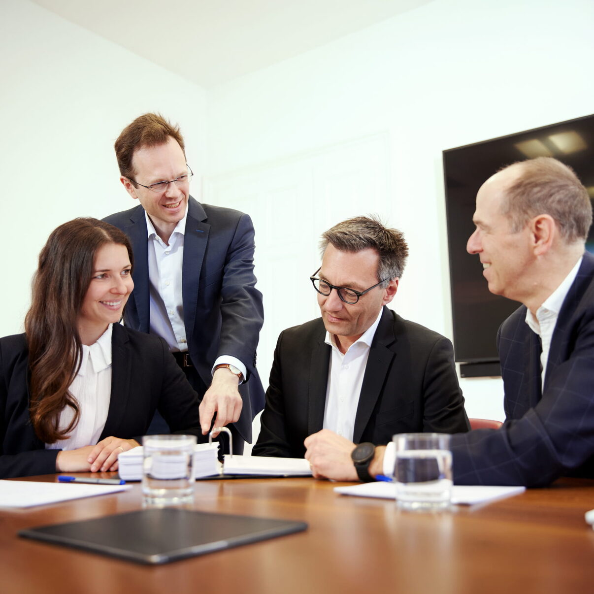 drei Personen sitzen an einem Tisch, eine steht dahinter und zeigt auf ein Dokument