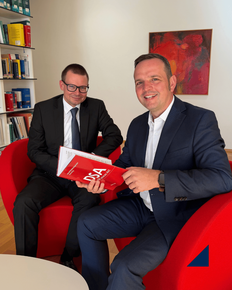 Zwei Personen in Business-Kleidung sitzen in einem Besprechungsraum auf roten Sesseln. Eine Person hält den roten Großkommentar zum Digital Services Act (DSA) des MANZ Verlags in den Händen, während die zweite Person daneben sitzt. Im Hintergrund sind ein Bücherregal mit juristischer Fachliteratur sowie ein rot-orangefarbenes Gemälde zu sehen. Das Foto entstand in einer Kanzlei- bzw. Büroumgebung und verweist auf die Mitwirkung von Juristen am Großkommentar zum Digital Services Act.